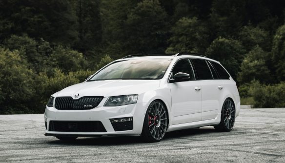 Fehér Škoda Octavia Combi áll egy parkolóban, zöld fák háttérrel.