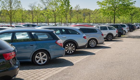 Parkoló autók sora, különböző színű és típusú járművekkel, zöld fákkal a háttérben.