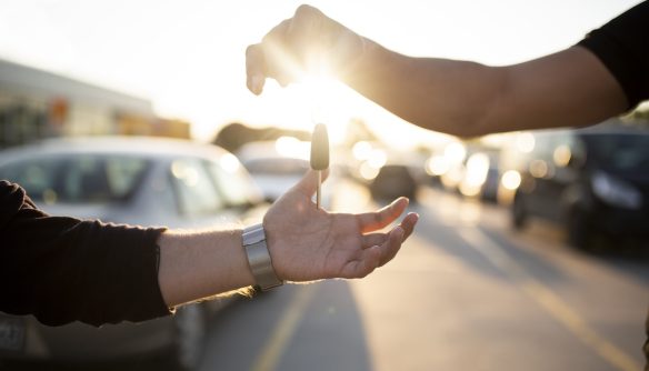 Két kéz, egy autókulcsot ad át az egyik a másiknak, háttérben parkoló autók és napfény.