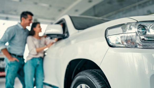 A képen egy fiatal pár látható, egy gépkocsit nézegetnek. A nő és a férfi mosolyogva vizsgálják a járművet, miközben közelről szemügyre veszik. A fókusz a jármű elején, különösen a fényszórón és az első keréken van, míg a pár kissé elmosódott a háttérben.