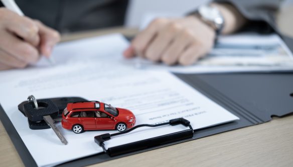 Gépjármű-átírás folyamatát illusztráló fotó. Egy ügyintéző papírokat ír alá, a kép fókuszában egy kisautó modell és kocsikulcs.