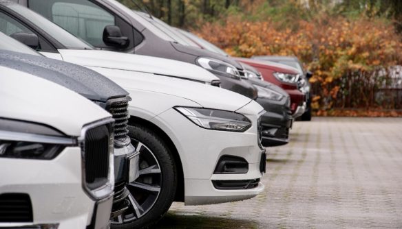 Többsoros parkolóban sorakozó különböző típusú személyautók – elsősorban SUV-ok és furgonok – esős, őszi időben. Az előtérben egy fehér Volvo, mellette többféle márka és színű használt jármű látható, háttérben színes lombú bokrokkal. A kép a használtautó-piac kínálatának sokszínűségét illusztrálja.