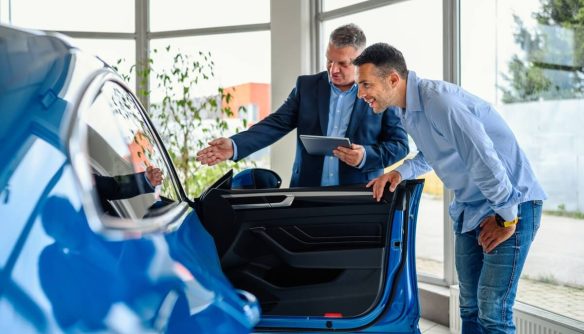 Autószalonban autót néző vásárló és értékesítő beszélgetnek egy kék jármű nyitott ajtaja mellett, táblagépen keresztül egyeztetve.