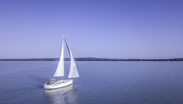 Egy fehér vitorlás hajó úszik a nyugodt, kék vizű Balatonon, napsütéses időben. A hajó vitorlái teljesen ki vannak bontva, a fedélzeten néhány ember napozik vagy pihen. A háttérben a tó túlpartja látható, ahol egy alacsony hegyvonulat és vízparti települések sejlenek fel. A tiszta égbolt és a végtelen víztükör békés, nyári hangulatot áraszt.