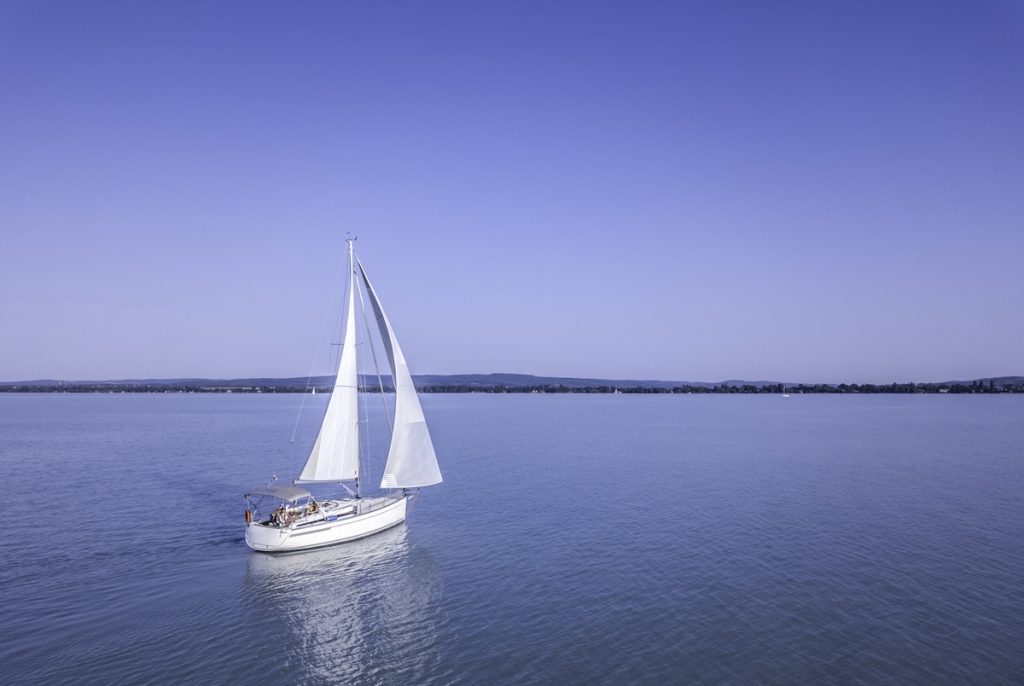 Egy fehér vitorlás hajó úszik a nyugodt, kék vizű Balatonon, napsütéses időben. A hajó vitorlái teljesen ki vannak bontva, a fedélzeten néhány ember napozik vagy pihen. A háttérben a tó túlpartja látható, ahol egy alacsony hegyvonulat és vízparti települések sejlenek fel. A tiszta égbolt és a végtelen víztükör békés, nyári hangulatot áraszt.