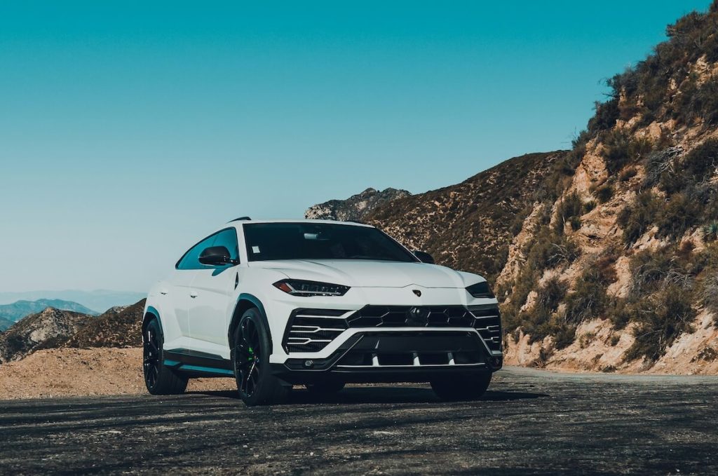 Fehér Lamborghini Urus terepjáró áll egy kanyargós hegyi úton, háttérben sziklás domboldallal és tiszta, kék égbolttal. A sportos megjelenésű luxus SUV a dinamizmust és az erőt sugározza természetes környezetben.
