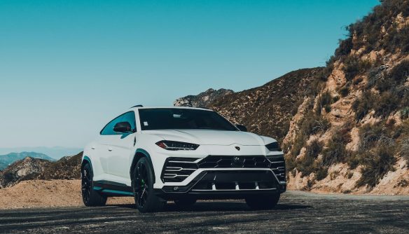 Fehér Lamborghini Urus terepjáró áll egy kanyargós hegyi úton, háttérben sziklás domboldallal és tiszta, kék égbolttal. A sportos megjelenésű luxus SUV a dinamizmust és az erőt sugározza természetes környezetben.