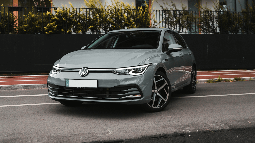 Szürke Volkswagen személyautó parkol egy városi utcán, modern fekete kerítéssel és bokrokkal a háttérben.