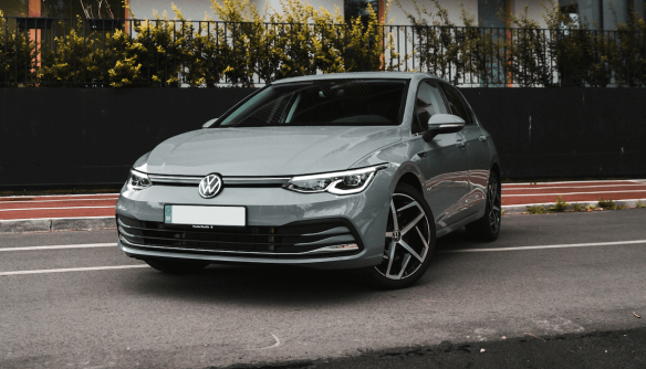 Szürke Volkswagen személyautó parkol egy városi utcán, modern fekete kerítéssel és bokrokkal a háttérben.