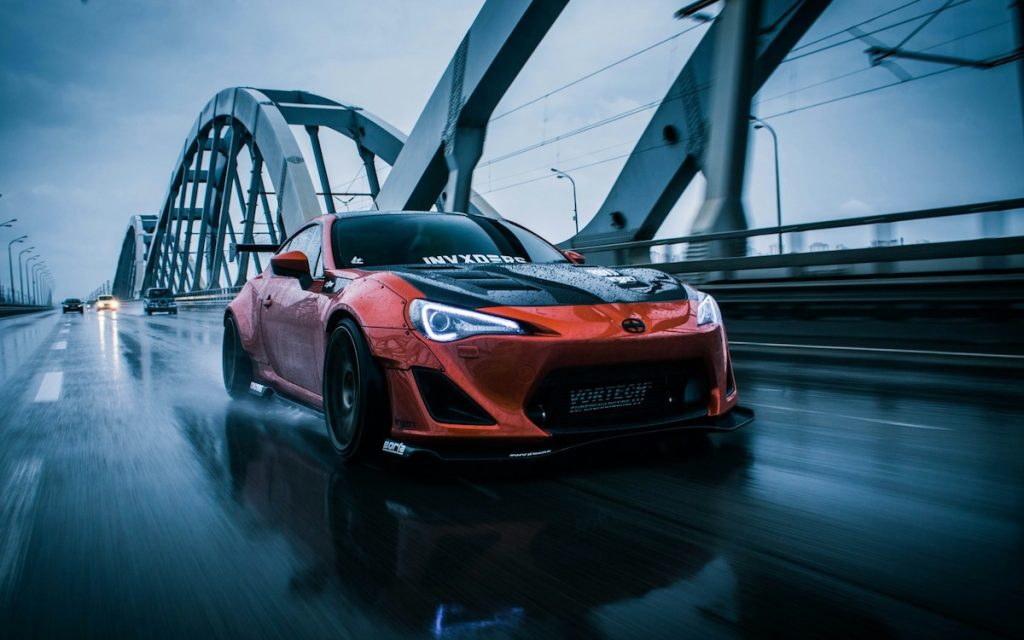 Piros Toyota sportautó esős úton, nagy sebességgel haladva egy modern acélhídon, LED fényszórókkal és sportos kialakítással.