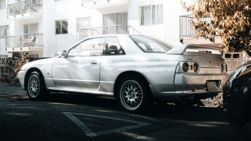 Egy fehér R32 GT-R oldalnézetből, ahogy egy út szélén parkol mögötte fehér lakóépület.