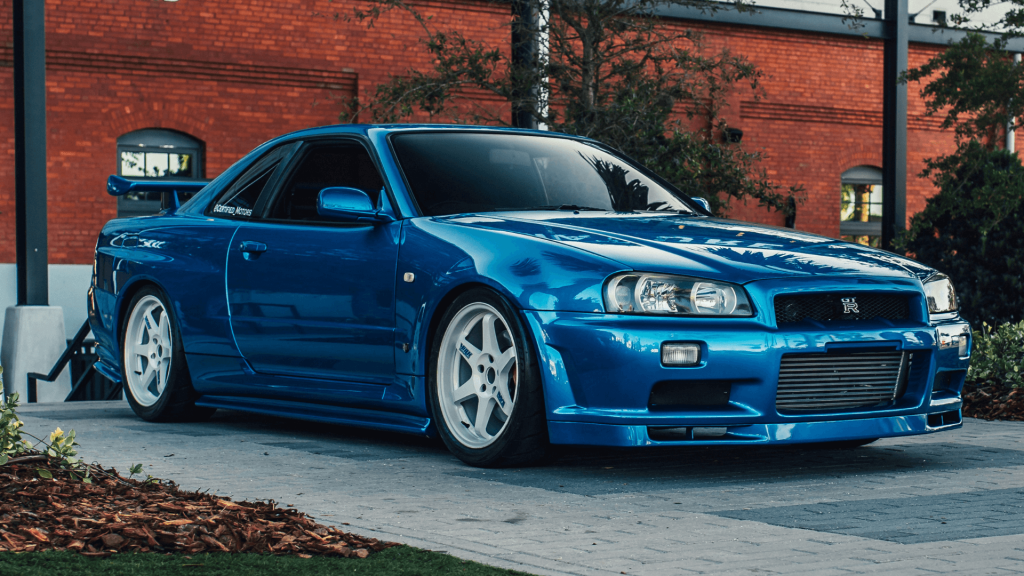 Ikonikus kék színű Nissan GT-R R34. A kép hátterében vörös téglafal és fák.