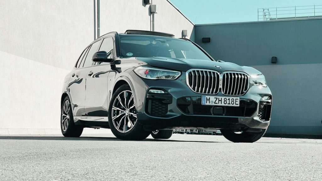 Fekete BMW SUV elölnézetből, napfényben, mögötte szürke beton épület.