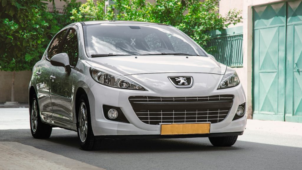 Fehér Peugeot személyautó parkol egy lakóövezet utcájában, zöld kapu és fák előtt.