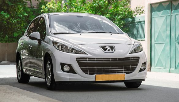 Fehér Peugeot személyautó parkol egy lakóövezet utcájában, zöld kapu és fák előtt.