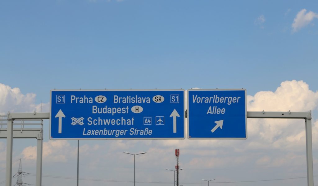 Autópálya irányjelző tábla, amely többek között Prága, Pozsony, Budapest és Schwechat felé mutat, jelezve a nemzetközi közúti kapcsolatokat és határátlépési útvonalakat. A kép a külföldi használt autó vásárlást és a gépjármű honosítást illusztrálja.