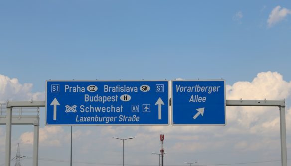 Autópálya irányjelző tábla, amely többek között Prága, Pozsony, Budapest és Schwechat felé mutat, jelezve a nemzetközi közúti kapcsolatokat és határátlépési útvonalakat. A kép a külföldi használt autó vásárlást és a gépjármű honosítást illusztrálja.