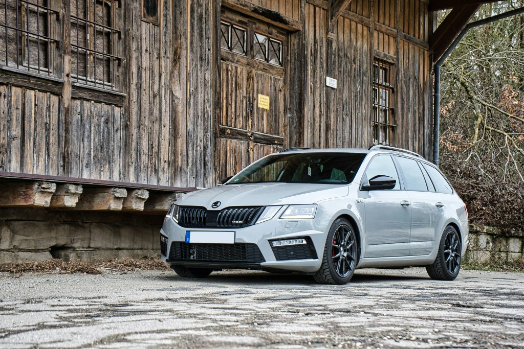 Szürke Škoda Octavia kombi parkol egy rusztikus, fából készült épület előtt. A modern autó fekete sportfelnikkel, piros féknyergekkel és sötétített ablakokkal látható, kontrasztban a természetes, vidéki háttérrel.