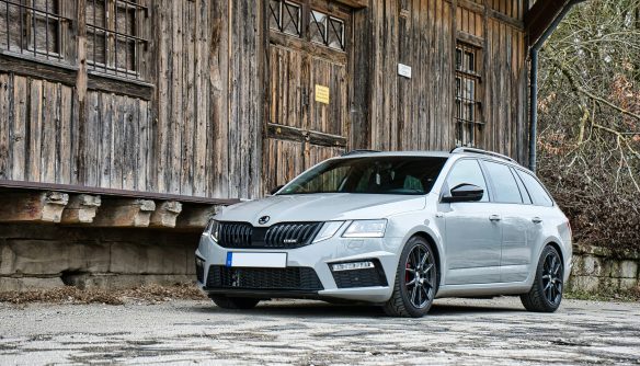 Szürke Škoda Octavia kombi parkol egy rusztikus, fából készült épület előtt. A modern autó fekete sportfelnikkel, piros féknyergekkel és sötétített ablakokkal látható, kontrasztban a természetes, vidéki háttérrel.