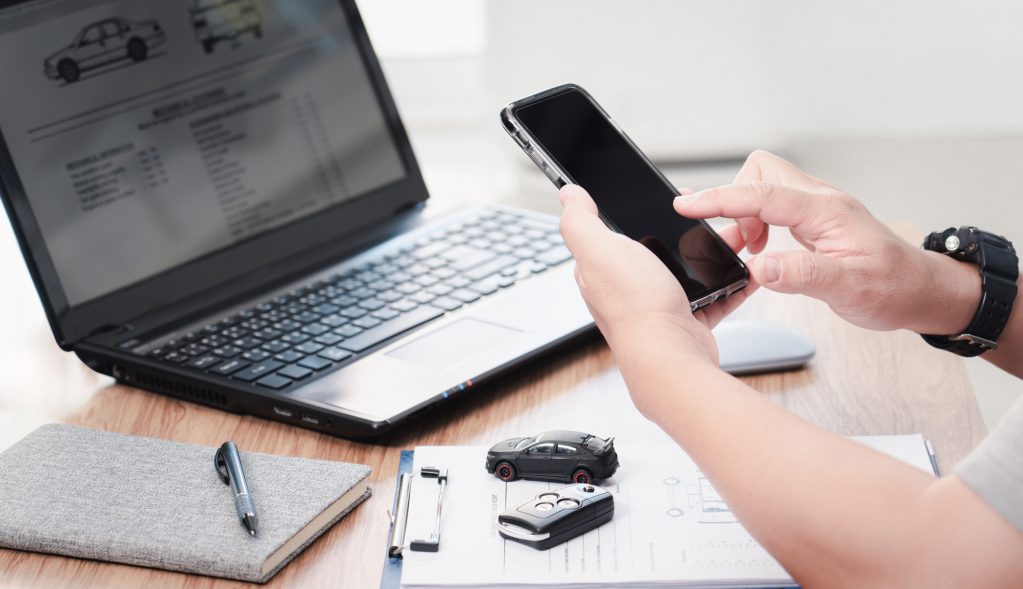 Egy ember okostelefont használ autóvásárlással kapcsolatos ügyintézés közben. Az asztalon laptop látható, amelyen autóadatlap jelenik meg, mellette jegyzetfüzet, toll, autókulcs és egy kisautó modell. A jelenet modern, digitális környezetet és online autópiaci tevékenységet ábrázol.