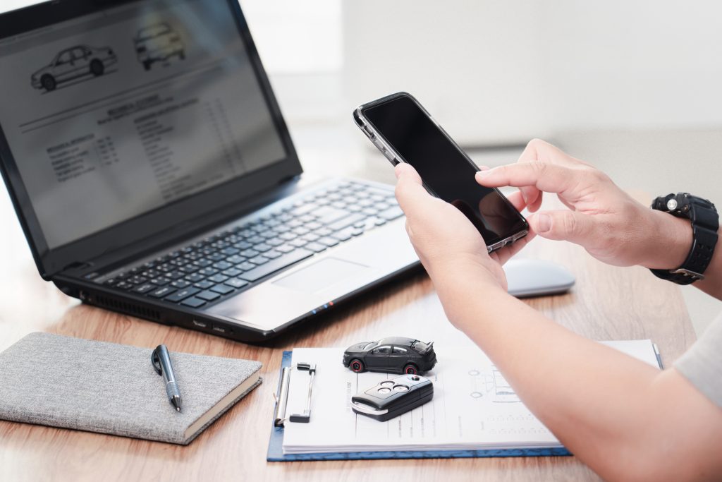 Egy ember okostelefont használ autóvásárlással kapcsolatos ügyintézés közben. Az asztalon laptop látható, amelyen autóadatlap jelenik meg, mellette jegyzetfüzet, toll, autókulcs és egy kisautó modell. A jelenet modern, digitális környezetet és online autópiaci tevékenységet ábrázol.