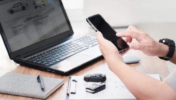 Egy ember okostelefont használ autóvásárlással kapcsolatos ügyintézés közben. Az asztalon laptop látható, amelyen autóadatlap jelenik meg, mellette jegyzetfüzet, toll, autókulcs és egy kisautó modell. A jelenet modern, digitális környezetet és online autópiaci tevékenységet ábrázol.
