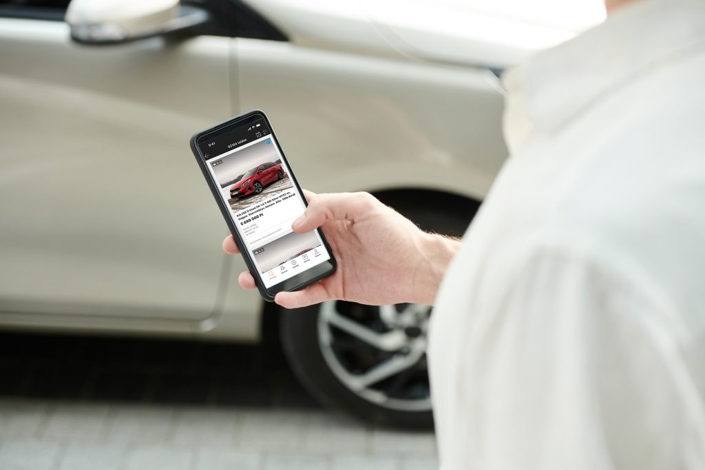 Egy férfi okostelefont tart a kezében, amin a Használtautó.hu applikáció látható, a háttérben pedig egy szürke autó.