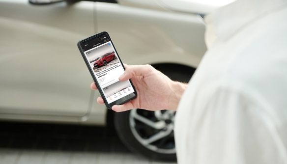 Egy férfi okostelefont tart a kezében, amin a Használtautó.hu applikáció látható, a háttérben pedig egy szürke autó.