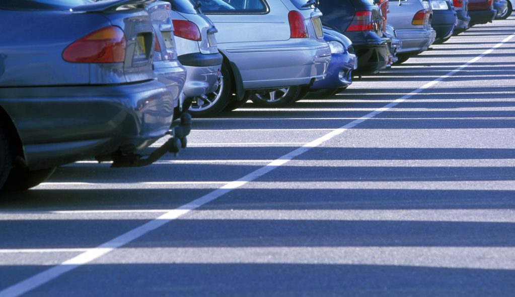 Parkolóban egymás mellett álló autók hátsó része látható, különböző színekben és típusokban. A kép alacsony szögből készült, a járművek sorai rendezett parkolóvonalak között húzódnak, erős napsütésben, hosszú árnyékokkal.