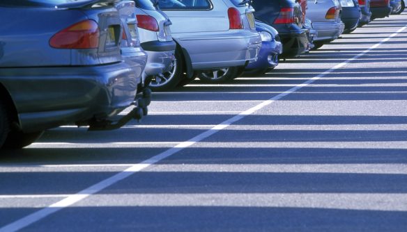 Parkolóban egymás mellett álló autók hátsó része látható, különböző színekben és típusokban. A kép alacsony szögből készült, a járművek sorai rendezett parkolóvonalak között húzódnak, erős napsütésben, hosszú árnyékokkal.