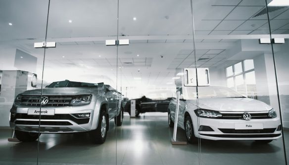 Egy szürke Volkswagen Amarok és egy fehér Polo a márkakereskedésben, a kirakat üvegén keresztül fotózva.
