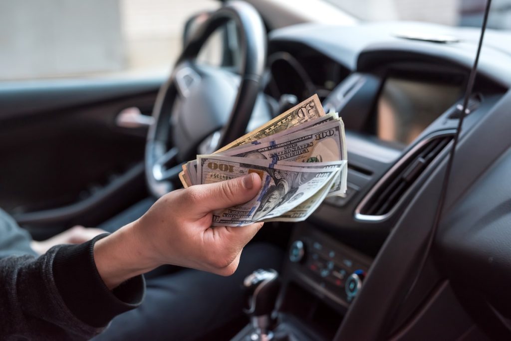 Egy férfi készpénzt nyújt a kamera felé egy autó sofőrüléséről.
