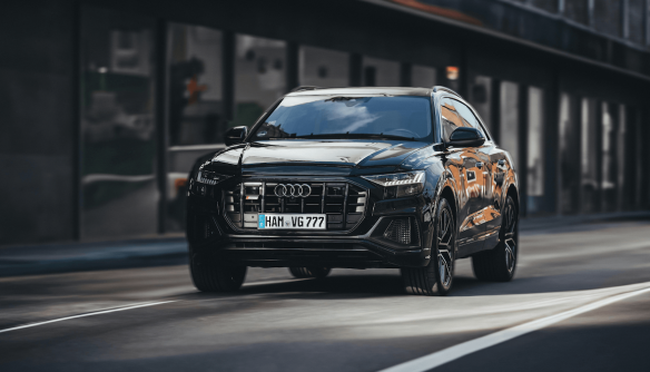 Fekete színű, modern Audi SUV halad egy városi utcán, elölről fotózva. A jármű fényes karosszériája tükrözi a környező épületeket, a háttér enyhén elmosódott, így a mozgás dinamikus hatást kelt.