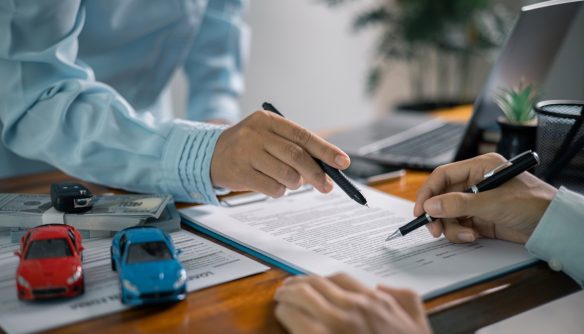 Autó eladó és vevő a gépjármű adásvételi szerződés aláírása közben. Az asztalon a szerződés mellett egy kék és egy piros játékautó látható.