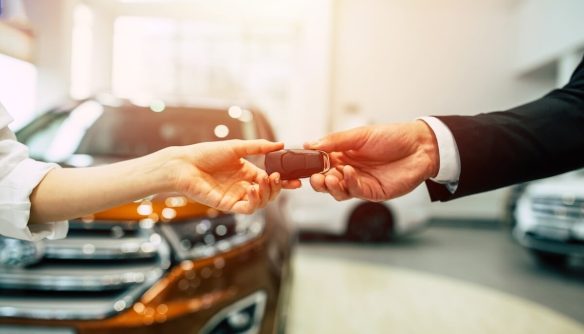 Az eladó átadja a kulcsot a használt autó vásárlónak. A háttérben egy autókereskedésben álló autó látható, amit megvilágít a napfény.