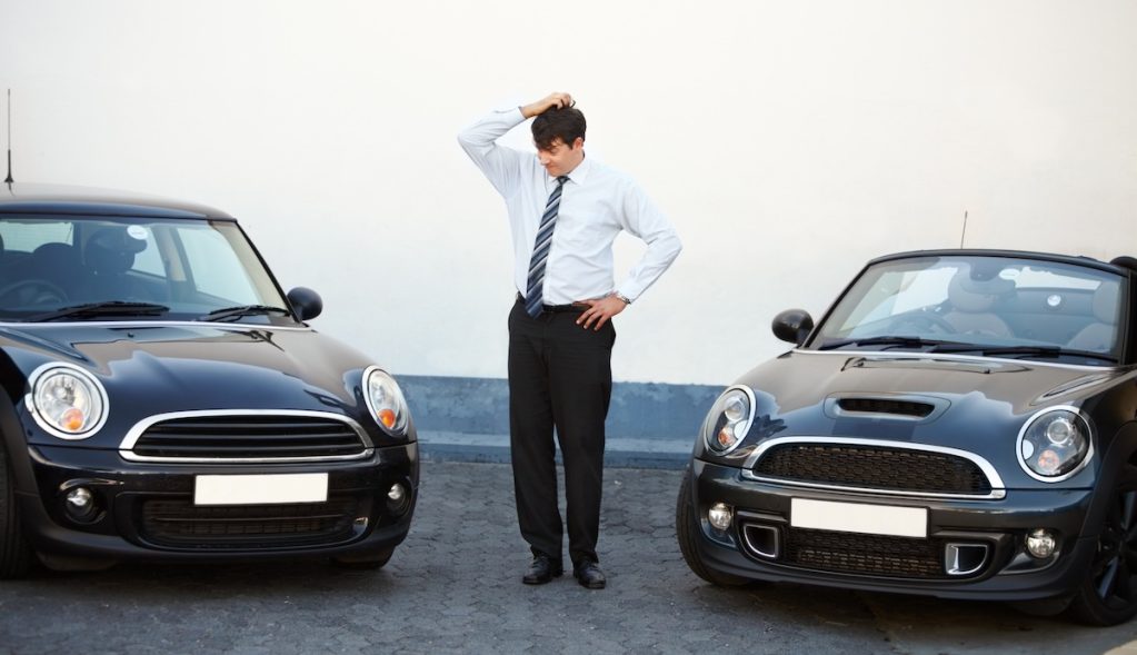 A képen két egyforma autó között áll egy elegánsan öltözött férfi, és a fejét vakarja. A kép azt fejezi ki, néha nehéz eldönteni a jónak tűnő hirdetések közül melyik akar átverni.