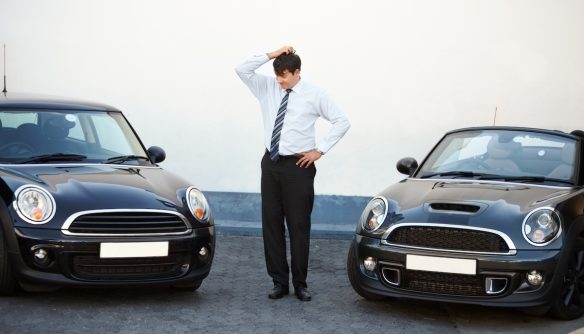 A képen két egyforma autó között áll egy elegánsan öltözött férfi, és a fejét vakarja. A kép azt fejezi ki, néha nehéz eldönteni a jónak tűnő hirdetések közül melyik akar átverni.