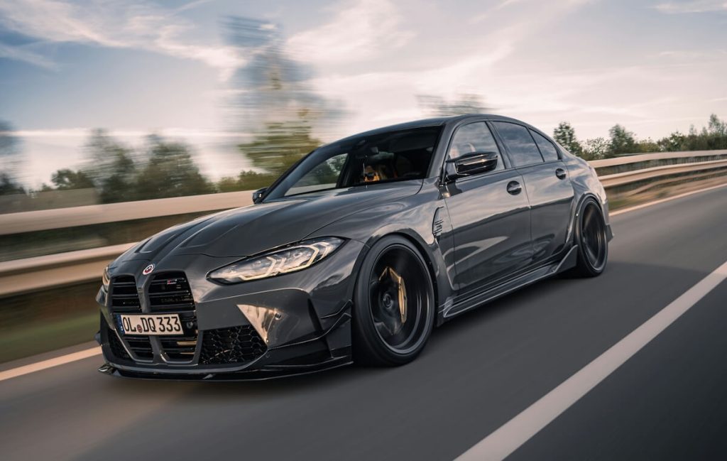 Sötétszürke BMW M3 hasít az autópályán, mögötte elmosódott fák és felhős ég.