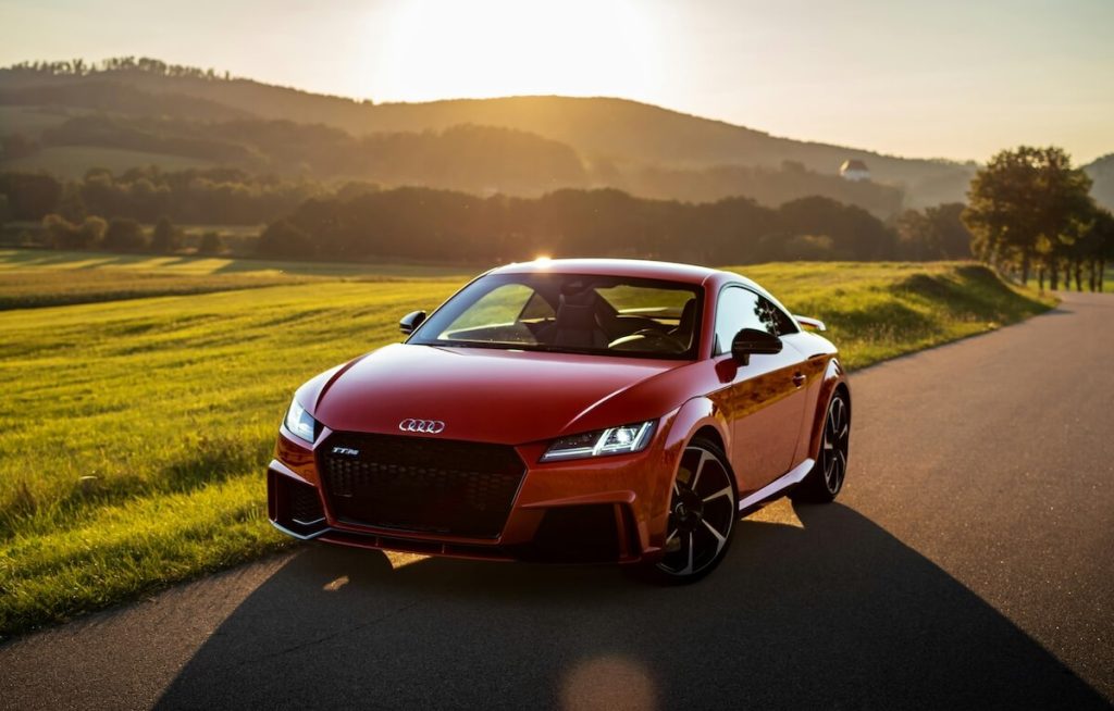 Piros Audi TT RS áll az út szélén a lemenő nap fényében, mögötte zöld mező és erdős dombok.