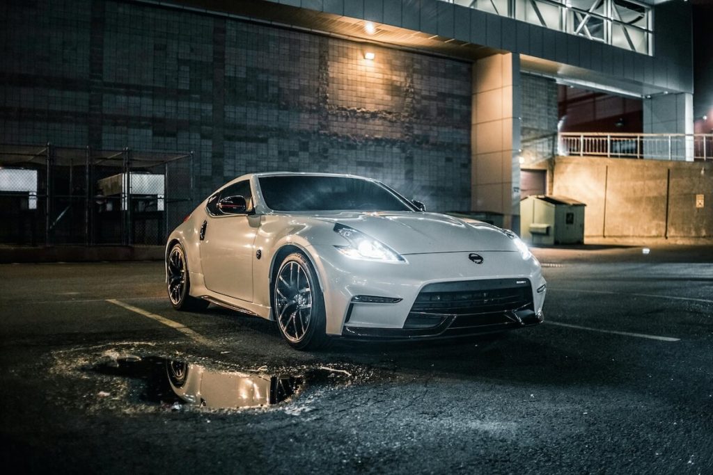 Fehér Nissan 370Z áll sötétedés után egy üres parkolóban, felkapcsolt lámpákkal. Mögötte egy szürke ipari épület. 
