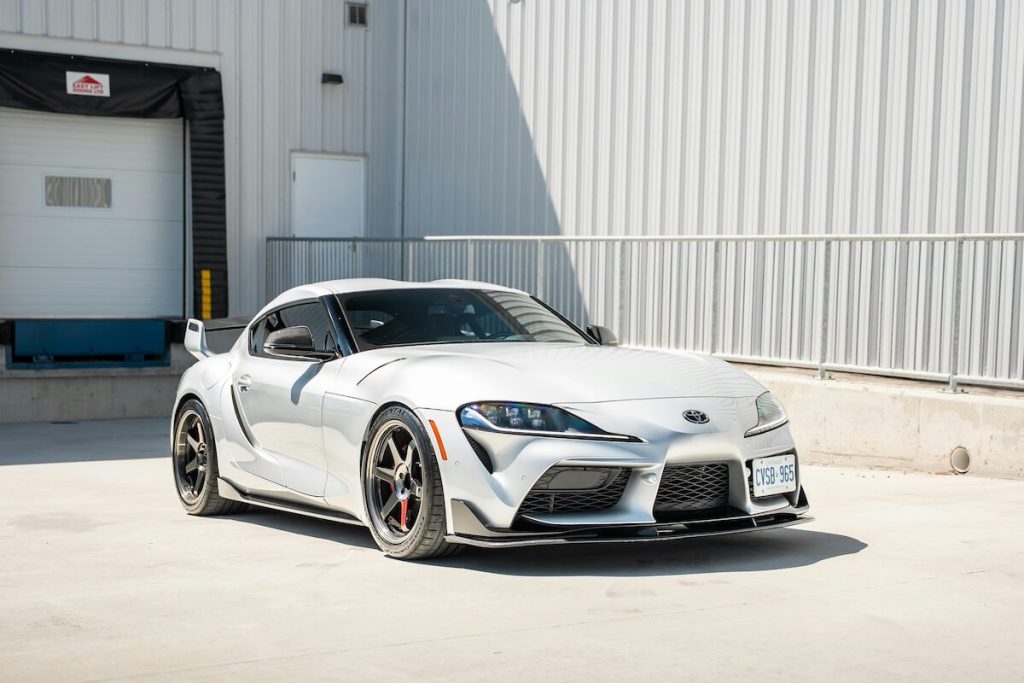 Világosszürke Toyota Supra verőfényes napsütésben, mögötte egy szürke ipari épülettel.