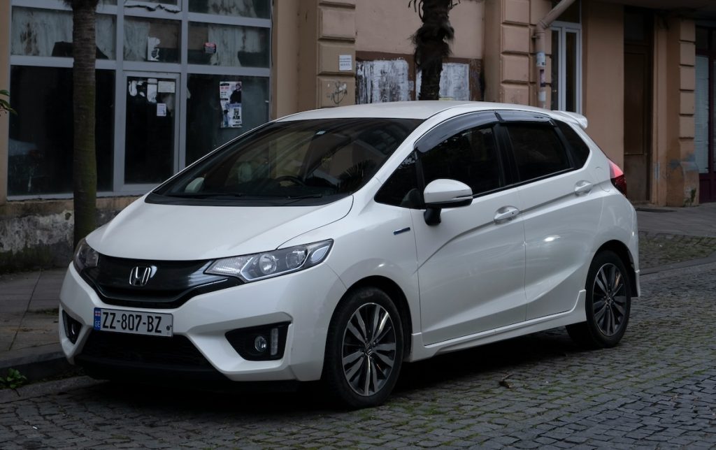Fehér Honda Jazz parkol az út szélén, mögötte épületek ablakai.