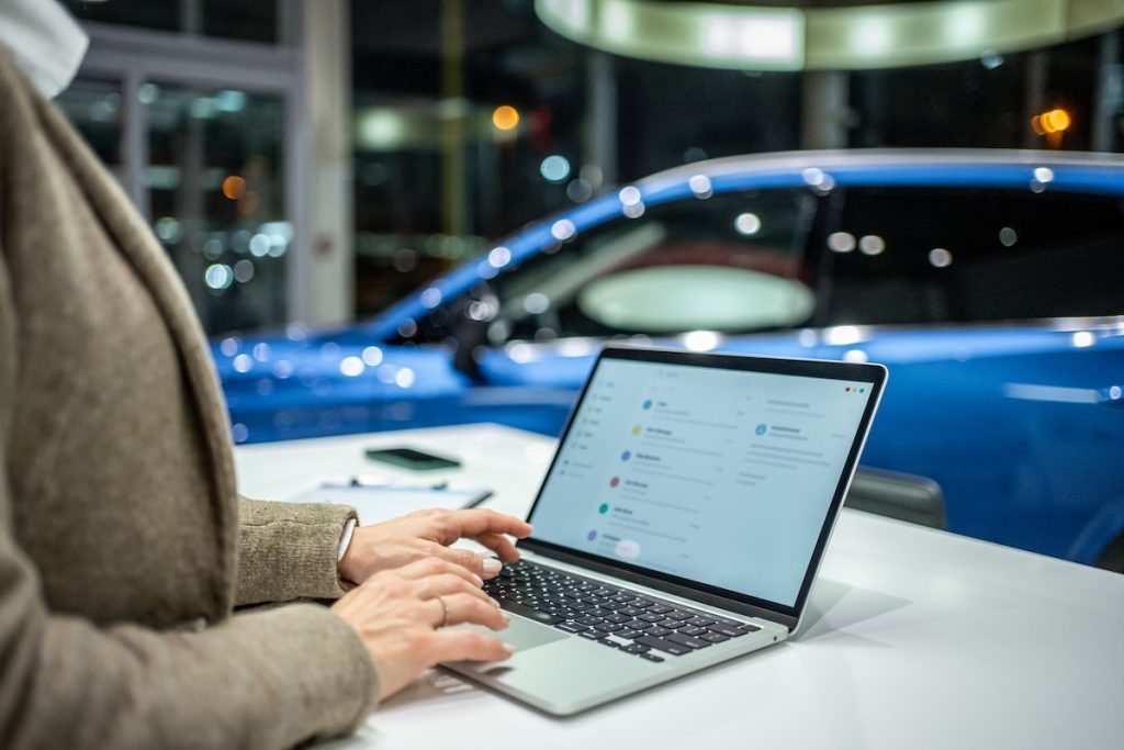 Eladó az autókereskedésben a laptopján dolgozik. A háttérben egy eladó kék használt autó látszik, elmosódva.
