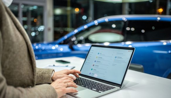 Eladó az autókereskedésben a laptopján dolgozik. A háttérben egy eladó kék használt autó látszik, elmosódva.