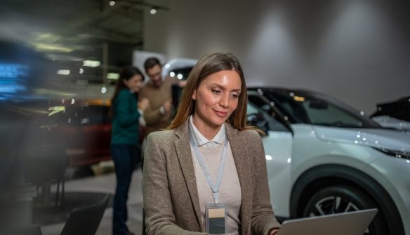 Autókereskedő hölgy laptopon tölti dolgozik a hirdetésfeltöltésen, mögötte eladó autók és érdeklődők.