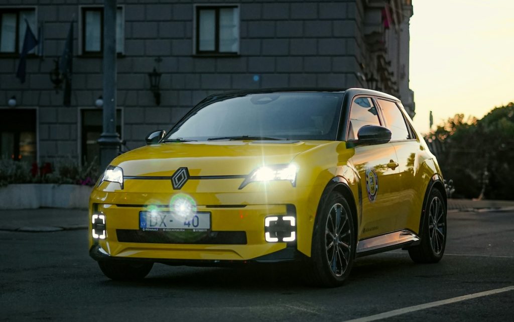 Sárga elektromos Renault 5 felkapcsolt lámpákkal a sötét városi utcákon