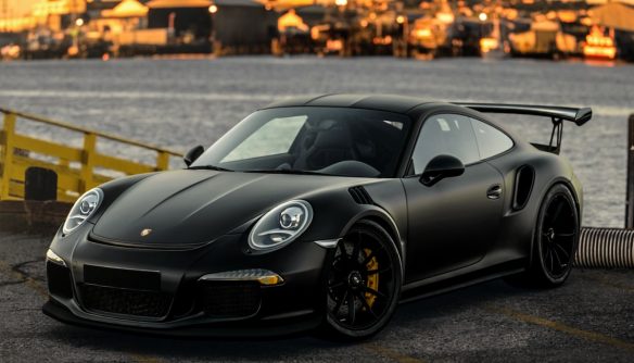 Fekete Porsche 911 GR3RS parkol a vízparton, mögötte naplemente és kikötő