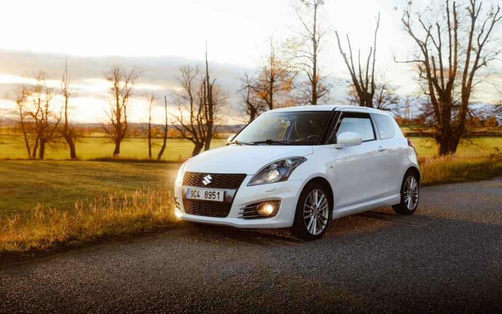 Fehér Suzuki Swift parkol az út szélén, mögötte naplemente.