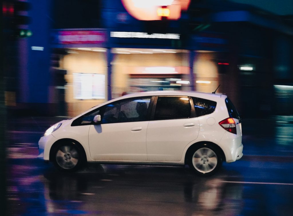 Fehér Honda Jazz sötétedés után az úton, mögötte városi fények elmosódva