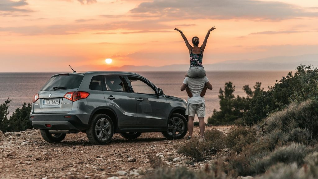Szürke Suzuki Vitara hátulról fotózva, tengerparti panoráma és naplemente előtt, mellette egy nő ül a párja nyakában és boldogan az ég felé emeli a kezeit.